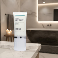 A white tube of Dr. Bailey Skin Care Daily Moisturizing Face Cream stands on a white marble bathroom counter with a blurred modern bathroom background featuring a bathtub and lit candles.