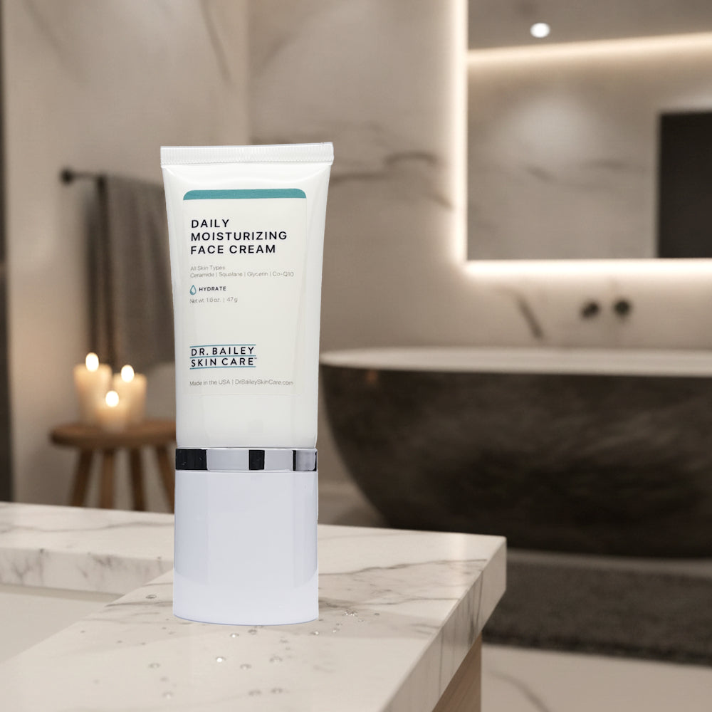 A white tube of Dr. Bailey Skin Care Daily Moisturizing Face Cream stands on a white marble bathroom counter with a blurred modern bathroom background featuring a bathtub and lit candles.